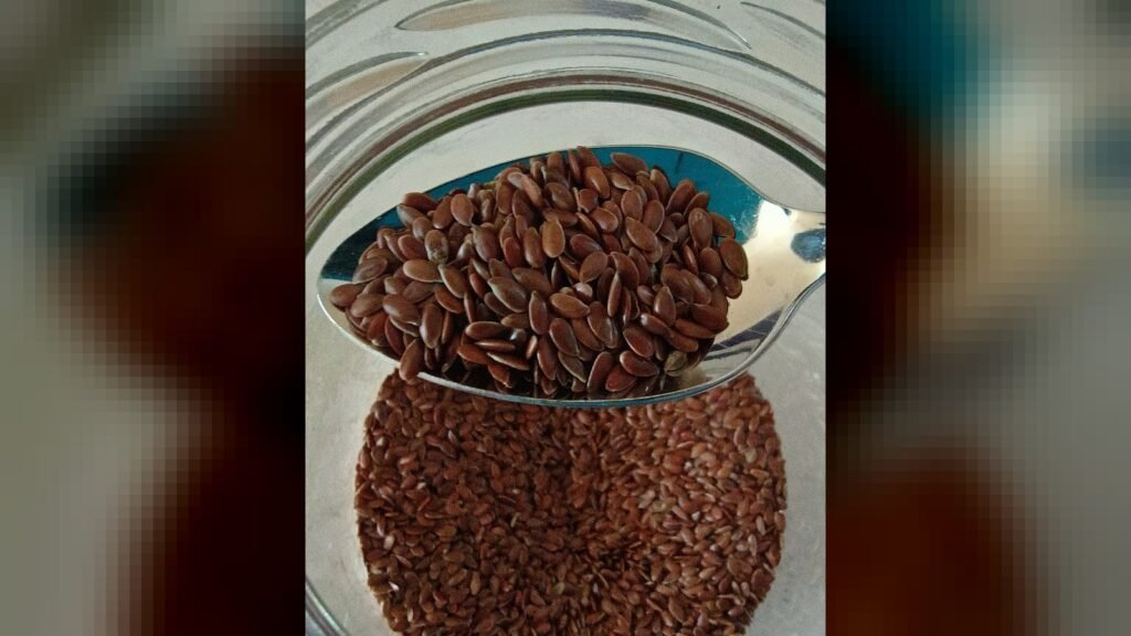 A spoonful of brown flaxseed is lifted out of a glass jar containing more flaxseed.