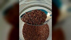 A spoonful of brown flaxseed is lifted out of a glass jar containing more flaxseed.