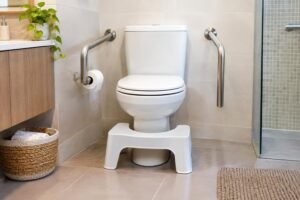 Banheiro com vaso sanitário branco, banquinho branco para apoiar os pés em frente ao vaso e barras de apoio metálicas fixadas nas paredes ao lado do vaso. Ao fundo, pia com vaso de planta, cesta com toalhas e box de vidro.