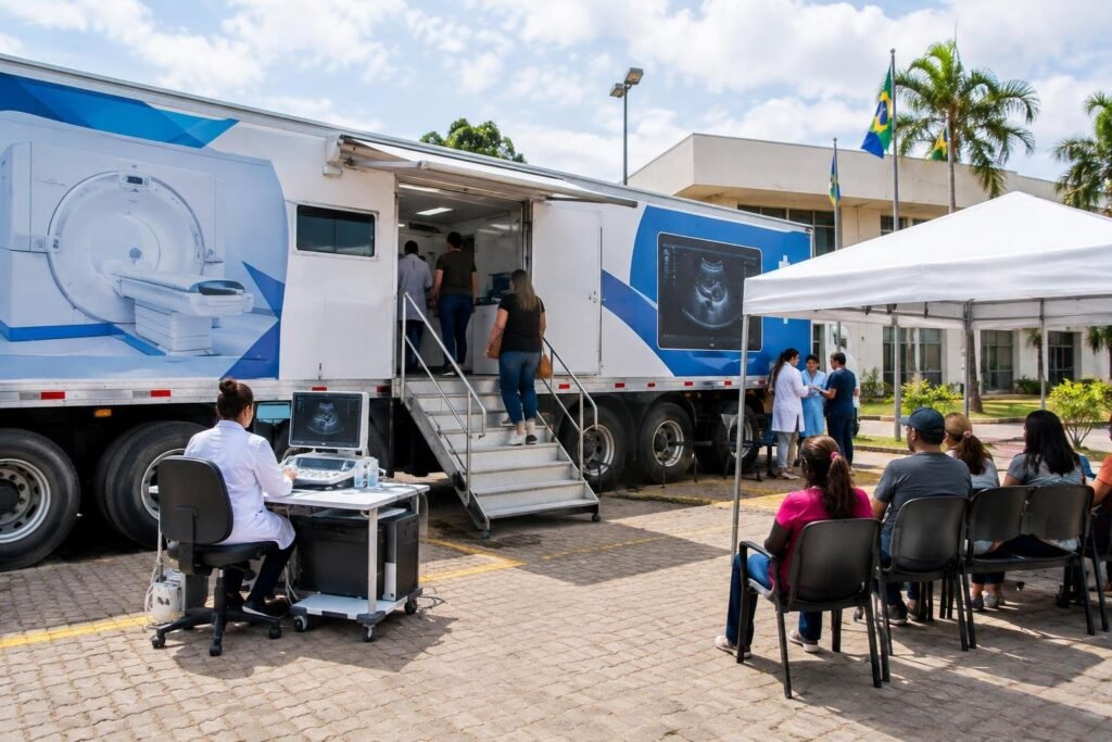 Carreta do Ministério da Saúde para exames de tomografia e ultrassom estacionada no Paço Municipal de Vilhena. Pessoas entram na carreta pela escada. Profissional de saúde sentada em frente a computador médico fora da carreta. Alguns pacientes sentados em cadeiras sob tenda aguardam do lado de fora. Bandeiras do Brasil e outras ao fundo.